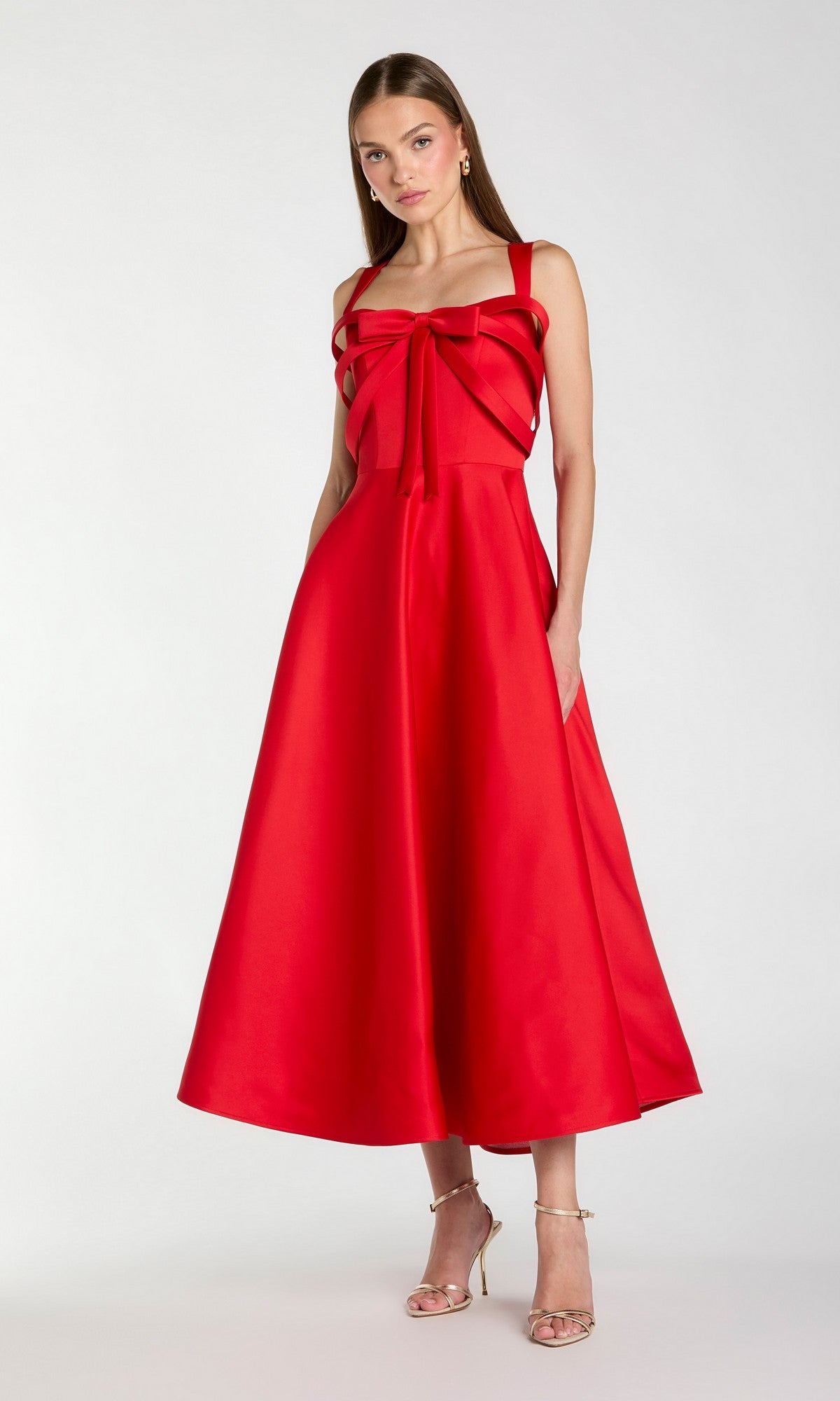A woman standing in a sleeveless, tea-length red Mac Duggal 21247 party dress with a large bow detail on the bodice, paired with gold high-heeled sandals.